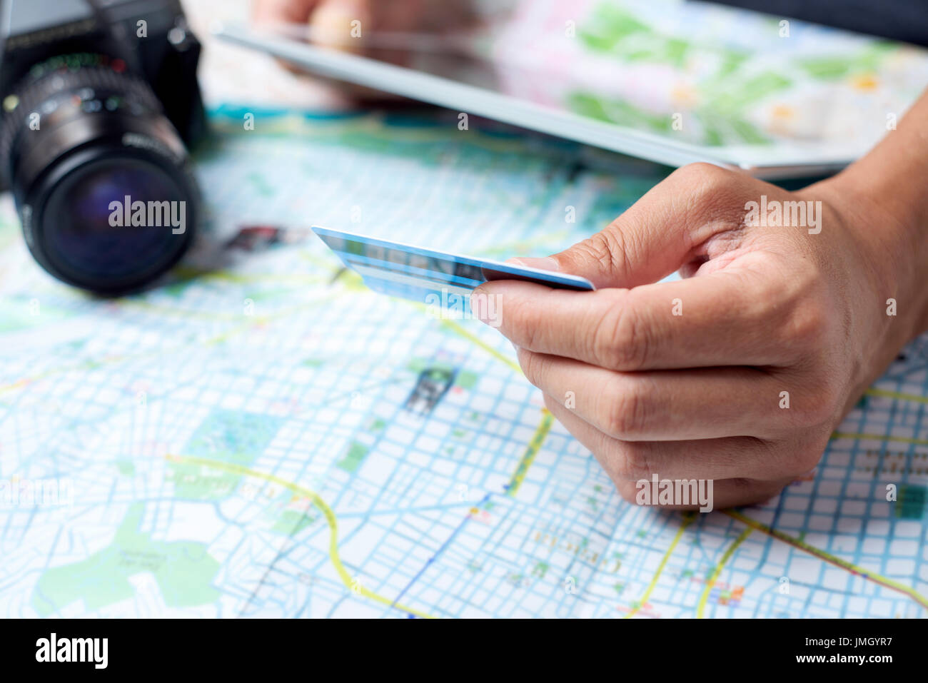 Primo piano di un giovane uomo caucasico utilizzando una carta di credito per prenotare un viaggio attraverso una tavoletta digitale, su un tavolo pieno di mappe e di una telecamera Foto Stock