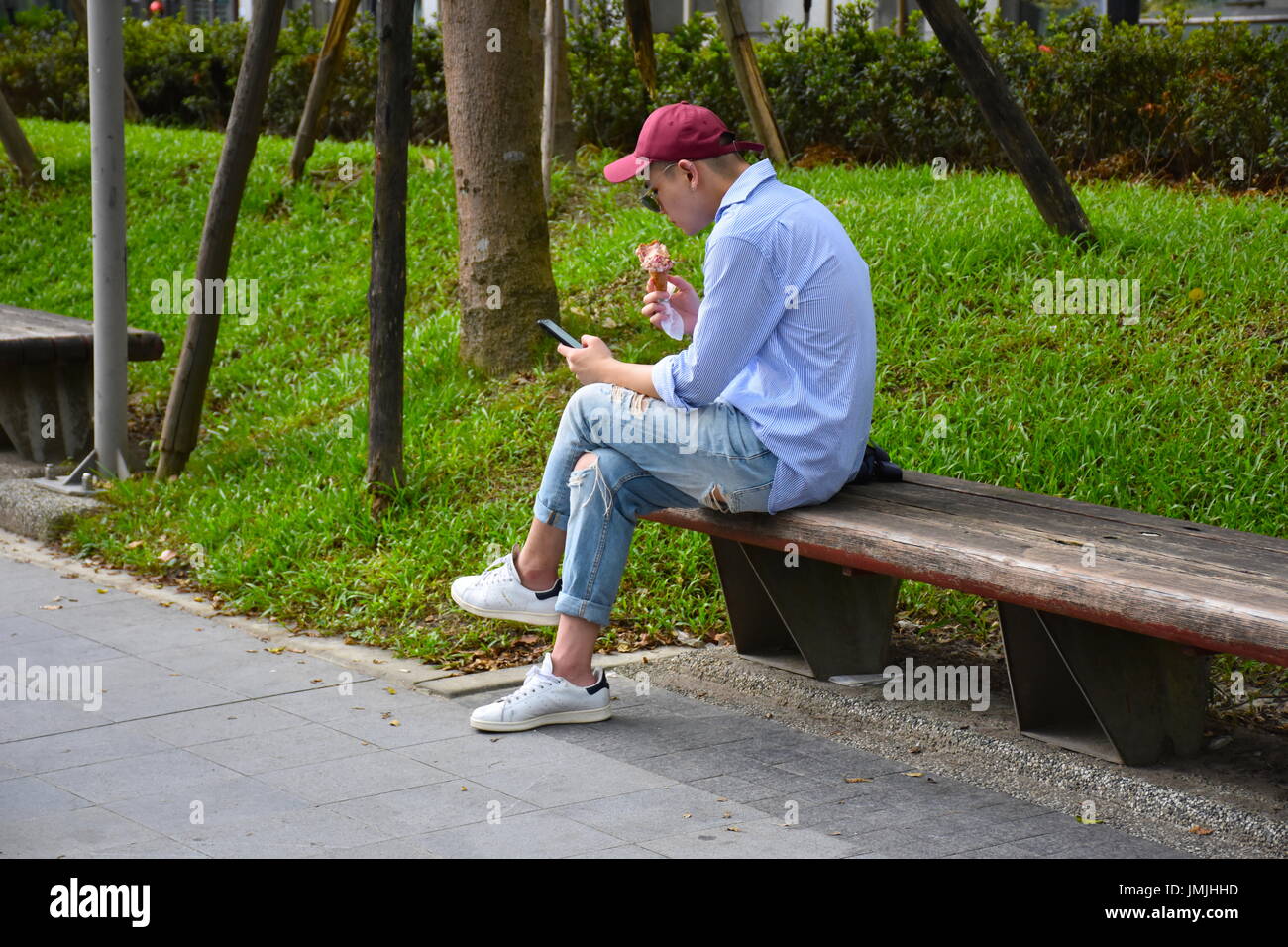 Un bel giovane uomo asiatico mangia un cono gelato mentre i messaggi di testo sul suo telefono, Taipei, Taiwan. Foto Stock