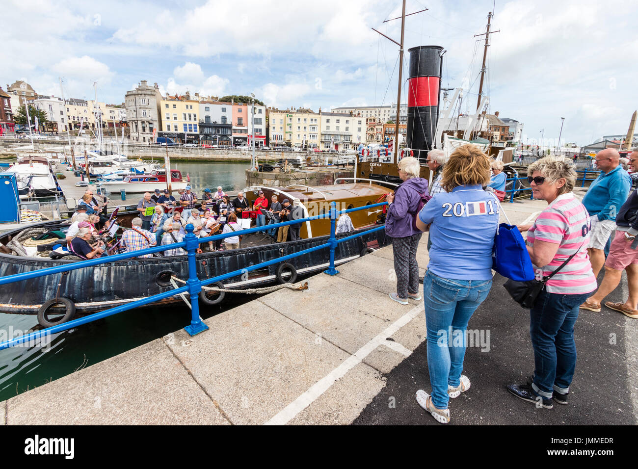 Concerto con la banda di trattativa, il Sunshine Ukes sul retro di un rimorchiatore a traino, il Cervia, ormeggiata in porto durante il festival di Ramsgate. Principalmente adulti senior, uomini e donne, seduto sul ponte di poppa della barca. Pubblico in piedi sulla banchina a guardare. Foto Stock