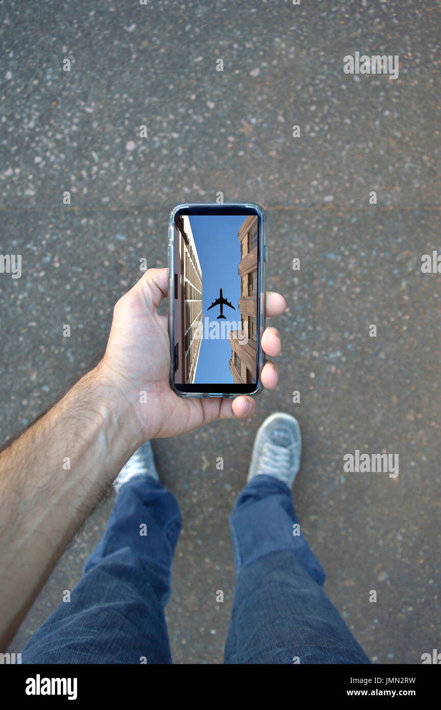 Parere personale di un uomo con il suo telefono cellulare in cui si è visto edifici dal livello basso ed un cielo blu chiaro con un aereo visto da sotto Foto Stock