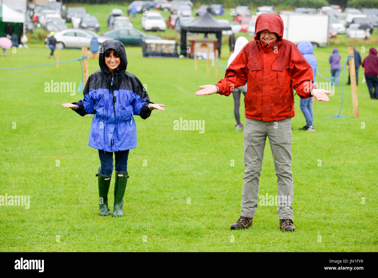 Coppia indossando impermeabili sotto la pioggia battente, spalle struggenti e sorridenti nell'estate inglese. Foto Stock