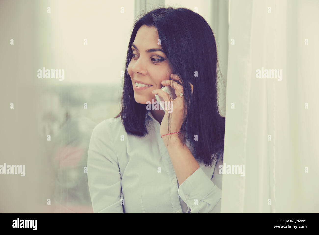 Giovane donna parlando al telefono cellulare mentre in piedi da una finestra in appartamento. Bellissimo modello femminile in Piano luminoso Foto Stock