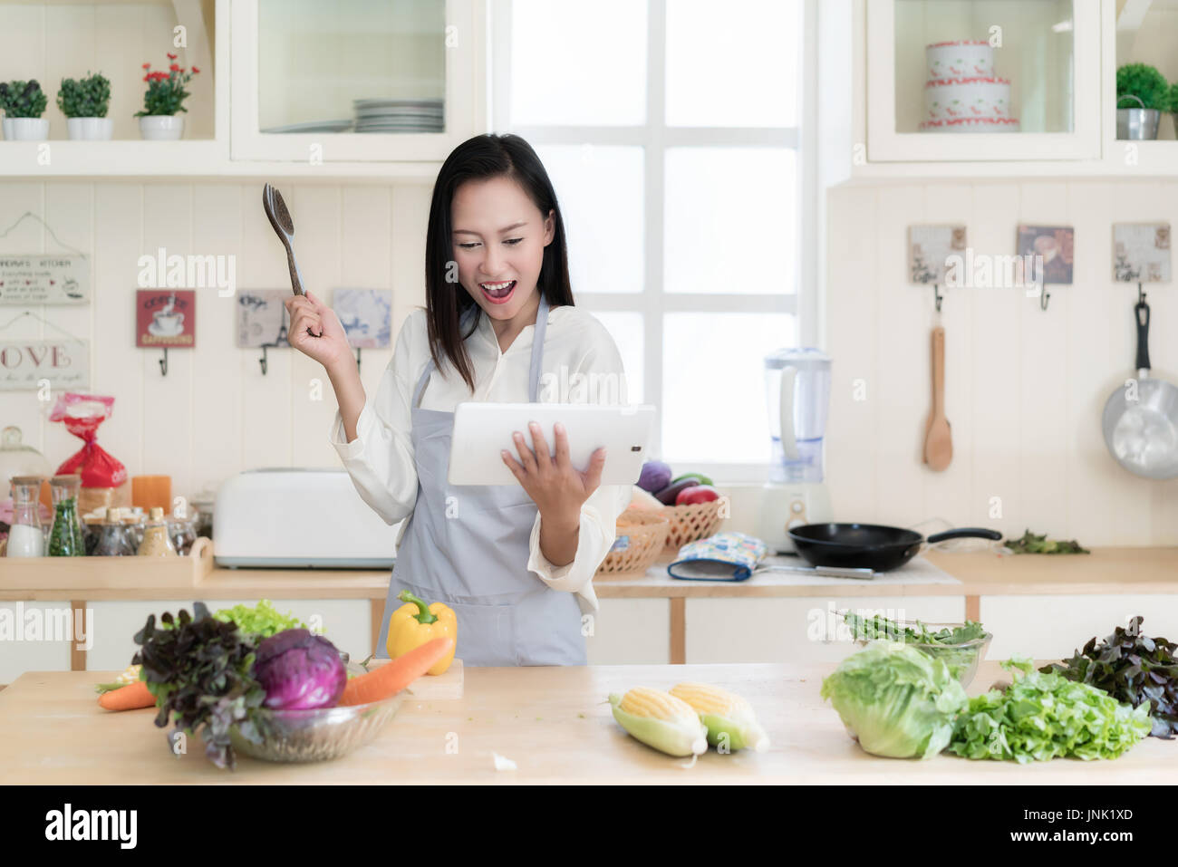 Allegro Asian giovane donna è la cucina in cucina con gioia. Ella è permanente e la holding digitale compressa della ricetta. Donna asiatica sta toccando un legno sp Foto Stock