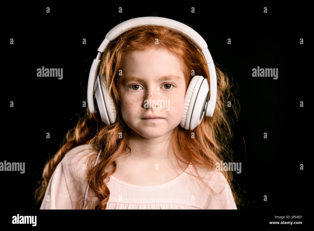 Ritratto di adorabile ragazza redhead ascolto di musica con cuffie guardando la telecamera isolato su nero Foto Stock