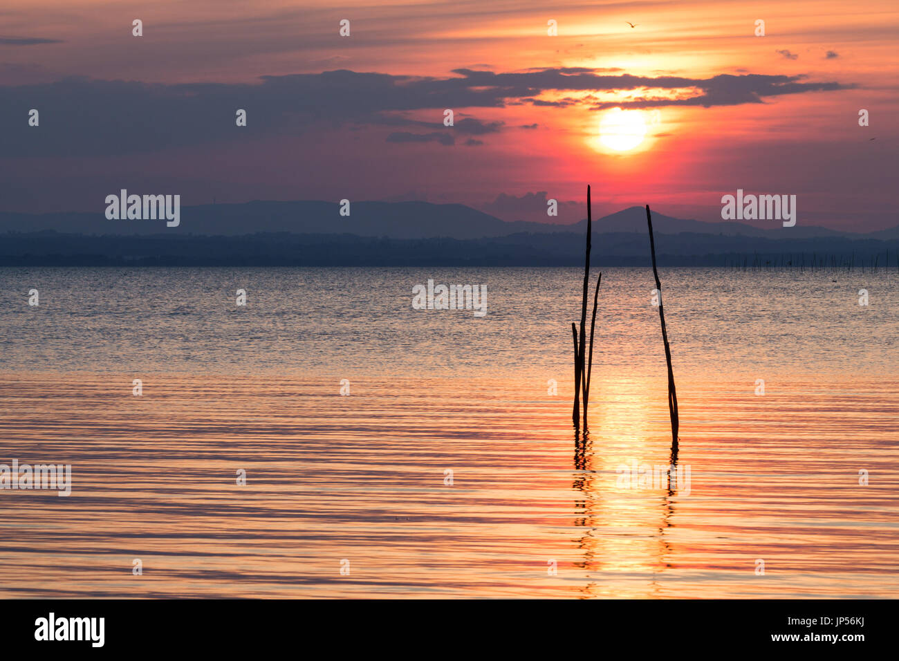 Tramonto al lago, con il sole basso sull'orizzonte, simmetrica riflessioni su acqua e alcuni pali in legno, con bellissimi colori tutto intorno Foto Stock