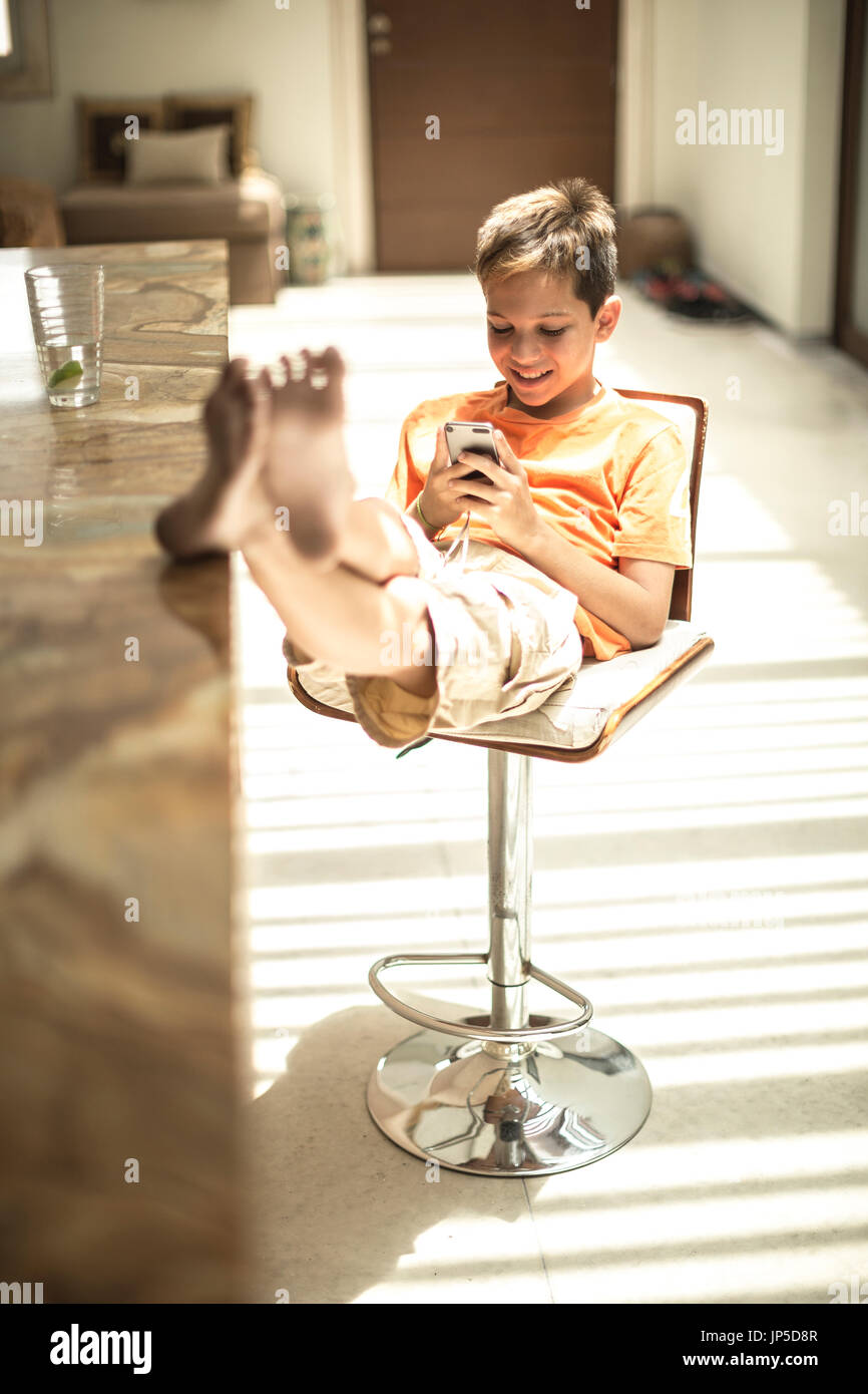 Un ragazzo seduto guardando un telefono cellulare schermo con i piedi su un banco di cucina. Foto Stock