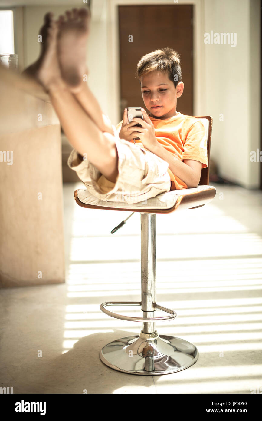 Un ragazzo seduto guardando un telefono cellulare schermo con i piedi su un banco di cucina. Foto Stock