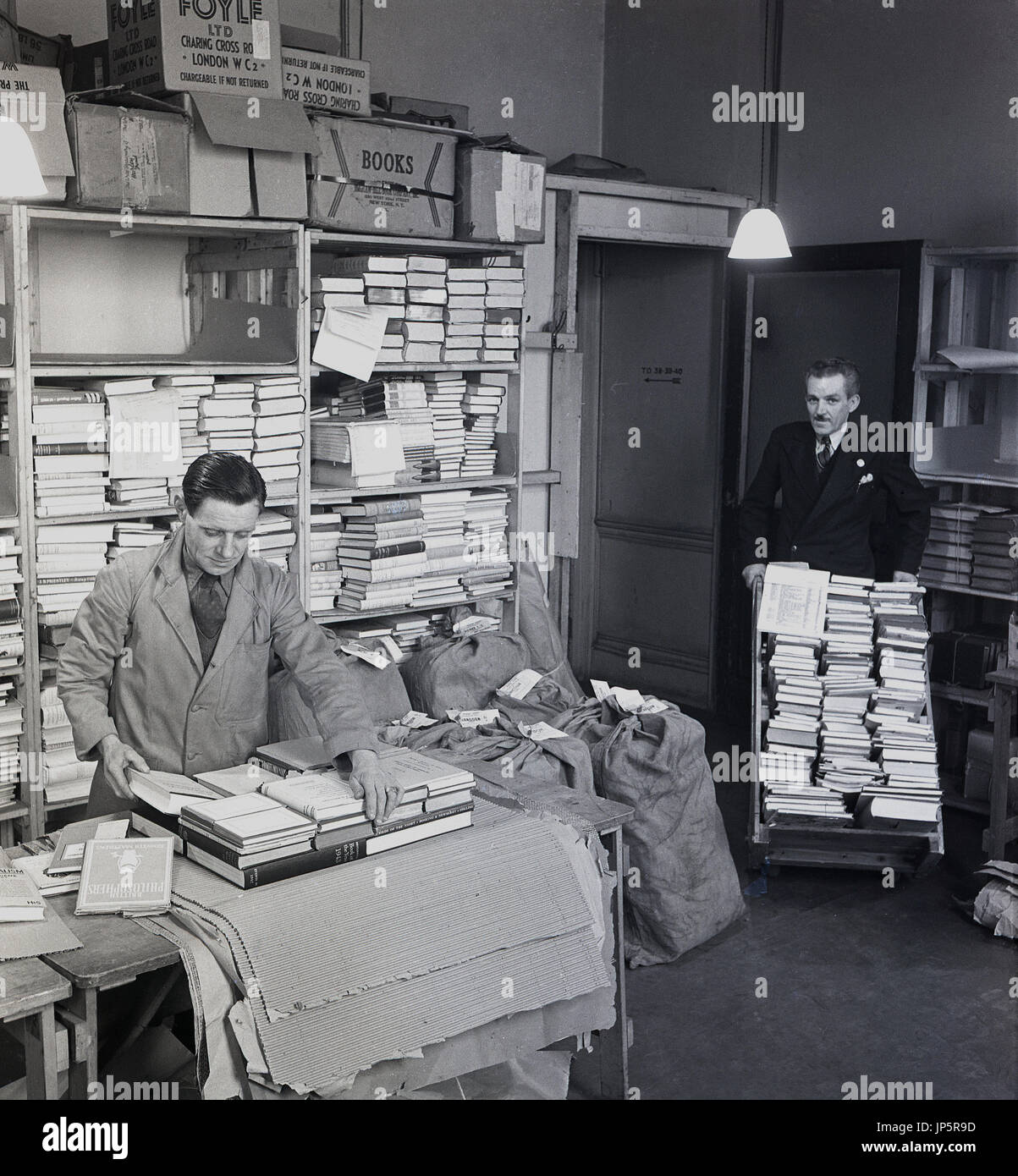 Degli anni Cinquanta, la figura mostra un dipendente rivestita con cravatta libri di imballaggio pronto per la spedizione in una camera di deposito presso il famoso bookshop di W & G Foyle Ltd, Charing Cross Rd, London, mentre un altro idoneo dipendente apporta più libri in su un carrello. Foto Stock