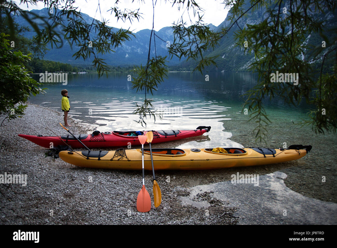 Giovane ragazzo in piedi sulla riva del tranquillo lago alpino da due kayak al crepuscolo. Il lago di Bohinj, Slovenia. Foto Stock