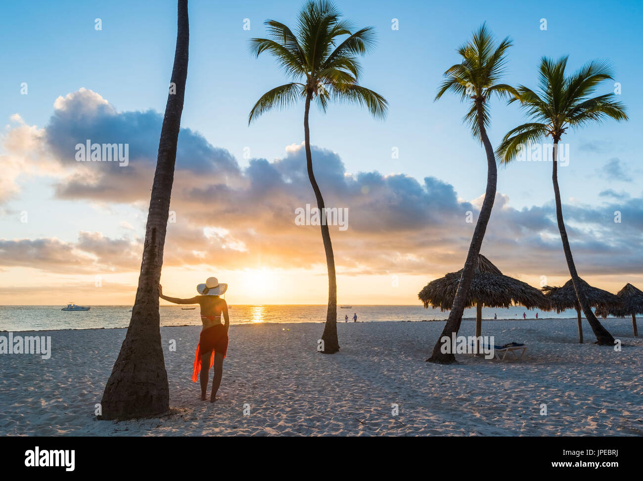 Bavaro Beach, Bavaro, Higuey, Punta Cana, Repubblica Dominicana. Donna ammirando il tramonto su una spiaggia orlata di palme (MR). Foto Stock