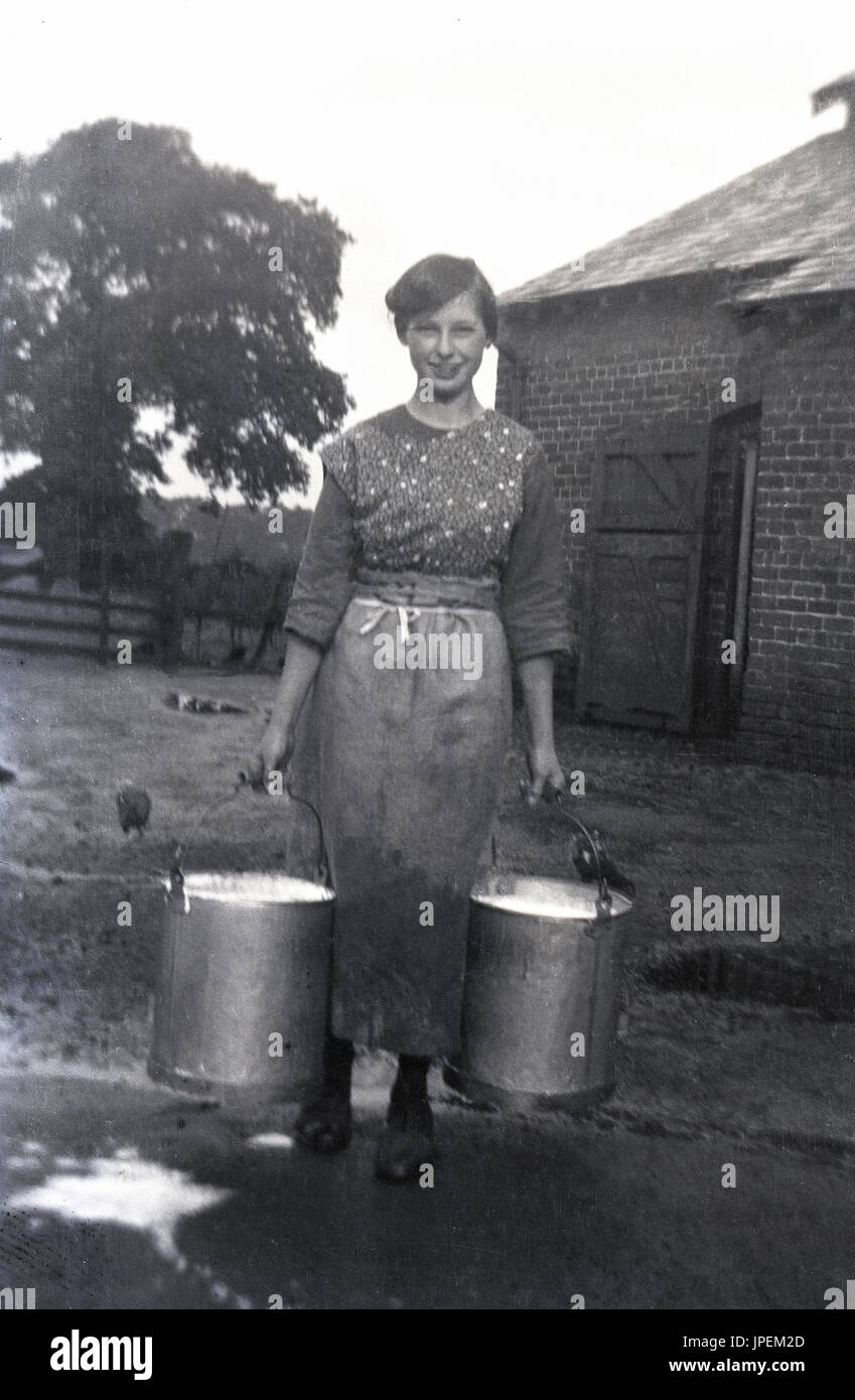 1930s, Inghilterra, immagine mostra un giovane agricoltore la moglie con grembiule al di fuori in giardino e porta due piena metai secchi del latte fresco. Foto Stock