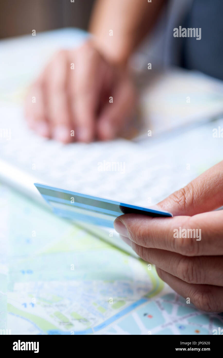 Primo piano di un giovane uomo caucasico utilizzando una carta di credito per prenotare un viaggio online utilizzando il suo computer su un tavolo pieno di mappe Foto Stock