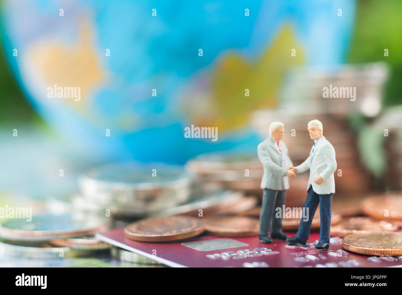 Imprenditore in miniatura e di handshaking permanente sulla carta di credito o carta di debito con globo sfocata e pile di monete sfondo usando come impegno, Foto Stock