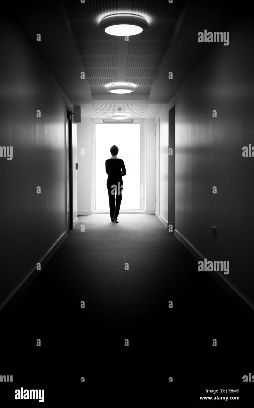 Donna in piedi da una finestra alla fine del corridoio, guardando fuori, simbolico "la luce alla fine del tunnel' vivere con la depressione. Foto Stock