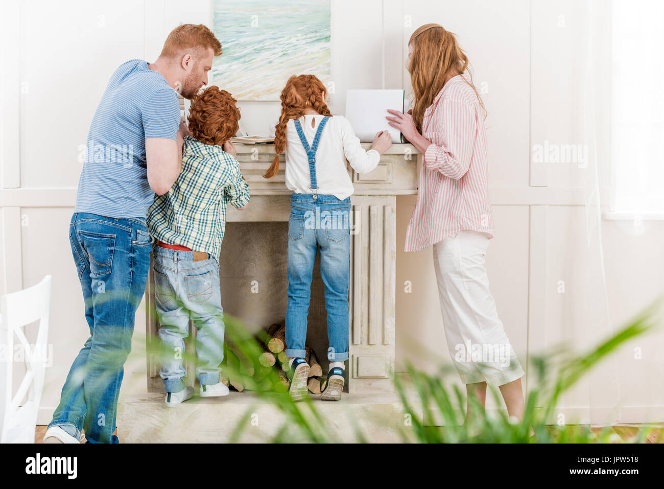 Vista posteriore del redhead famiglia con due bambini in piedi insieme vicino al camino e lettura di riviste, divertimento per tutta la famiglia al concetto di casa Foto Stock