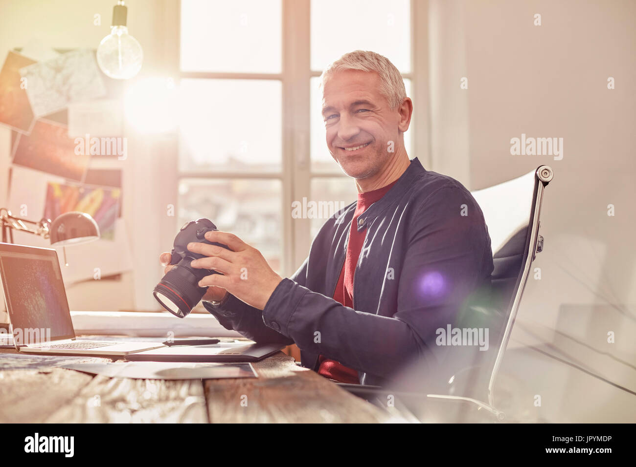 Ritratto sorridente e fiducioso fotografo maschio con fotocamera digitale lavora al computer portatile in ufficio soleggiato Foto Stock