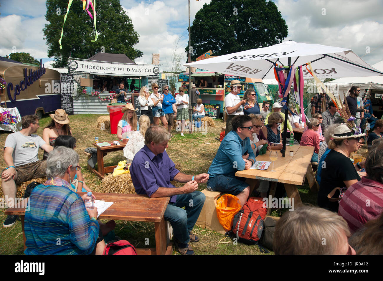 Famiglie godere il sole seduti a bere e mangiare fuori tende e tabelloni elettronici al porto Eliot Festival Cornwall Regno Unito Foto Stock