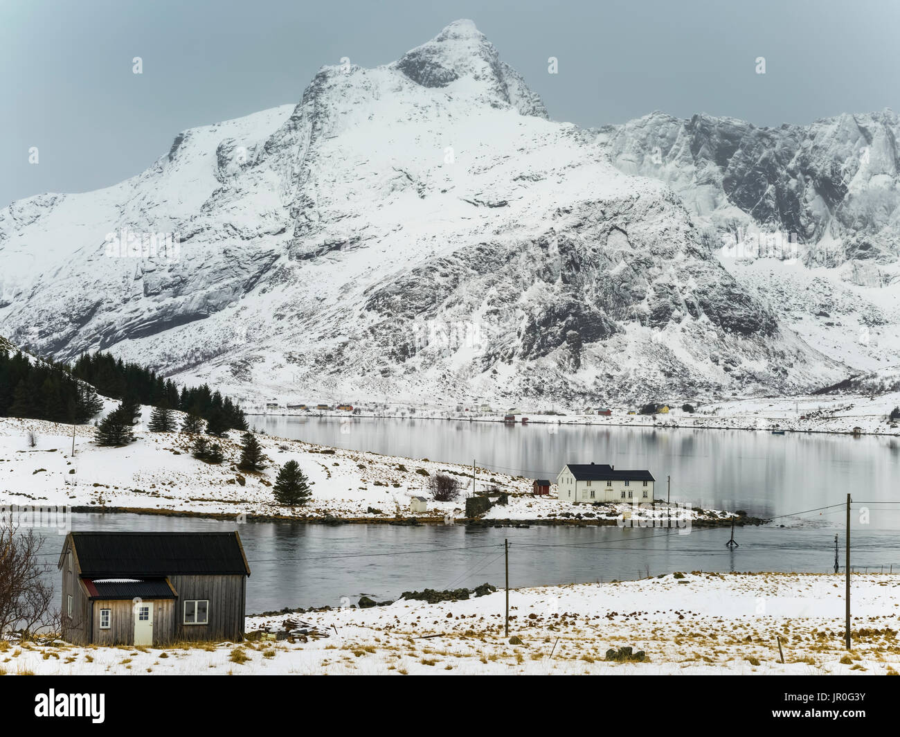 Robusto e il picco montagne coperte di neve e acqua tranquilla lungo la costa; Isole Lofoten, Nordland, Norvegia Foto Stock