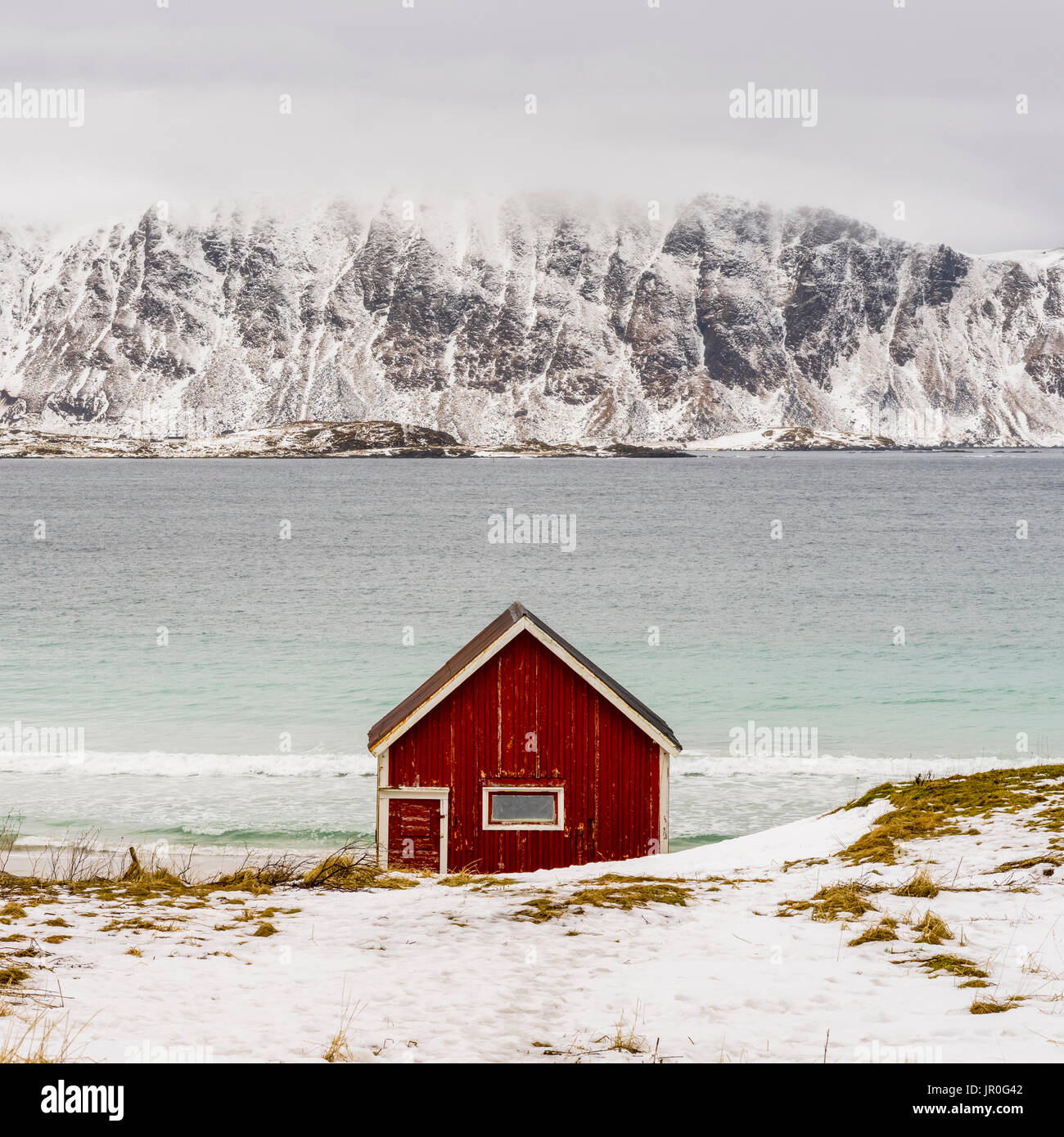 Una solitaria cabina rosso sul bordo dell'acqua con robusto e montagne coperte di neve in tutta l'acqua; Isole Lofoten, Nordland, Norvegia Foto Stock