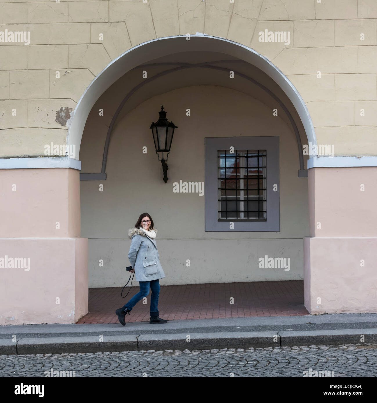Una donna passeggiate da un edificio sotto un arco con un sorriso, tenendo una telecamera dietro la schiena; Praga, Repubblica Ceca Foto Stock