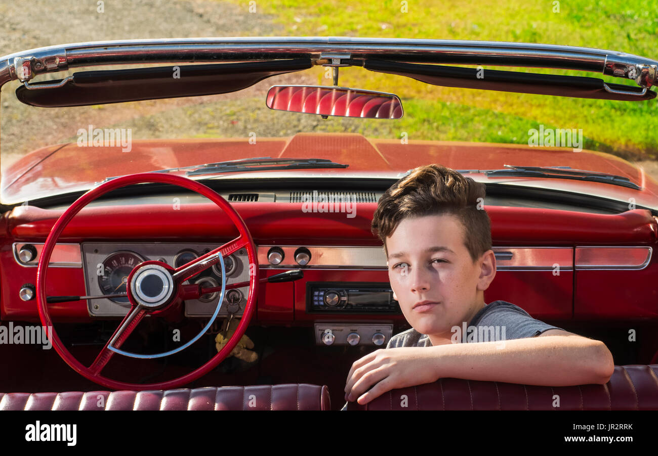 Ritratto di un Caucasian ragazzo adolescente con marrone capelli ondulati seduto con il suo braccio al di sopra del sedile di un 1960 Plymouth Valiant convertibile Foto Stock