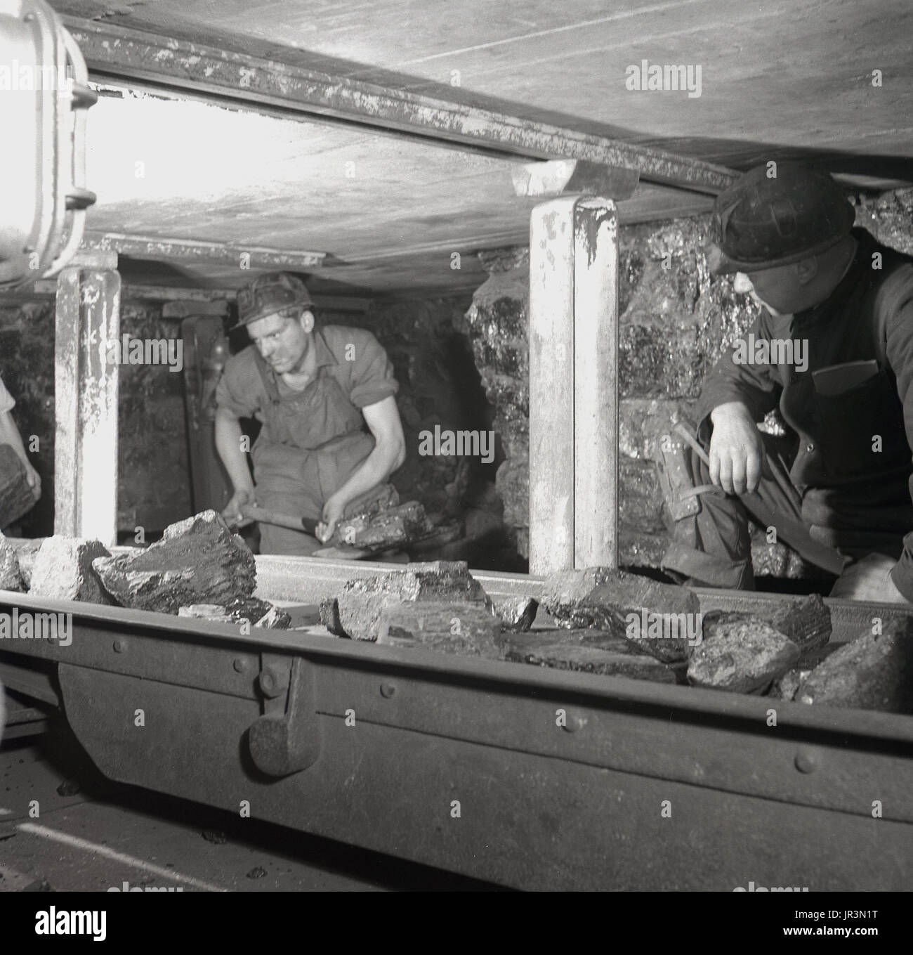 1948, storico Inghilterra, un minatore utilizzando una vanga per caricare grandi pezzi di carbone e di altre sostanze minerali in un trasportatore di metallo sotto l'occhio vigile di un esperto supervisore di data mining. Foto Stock