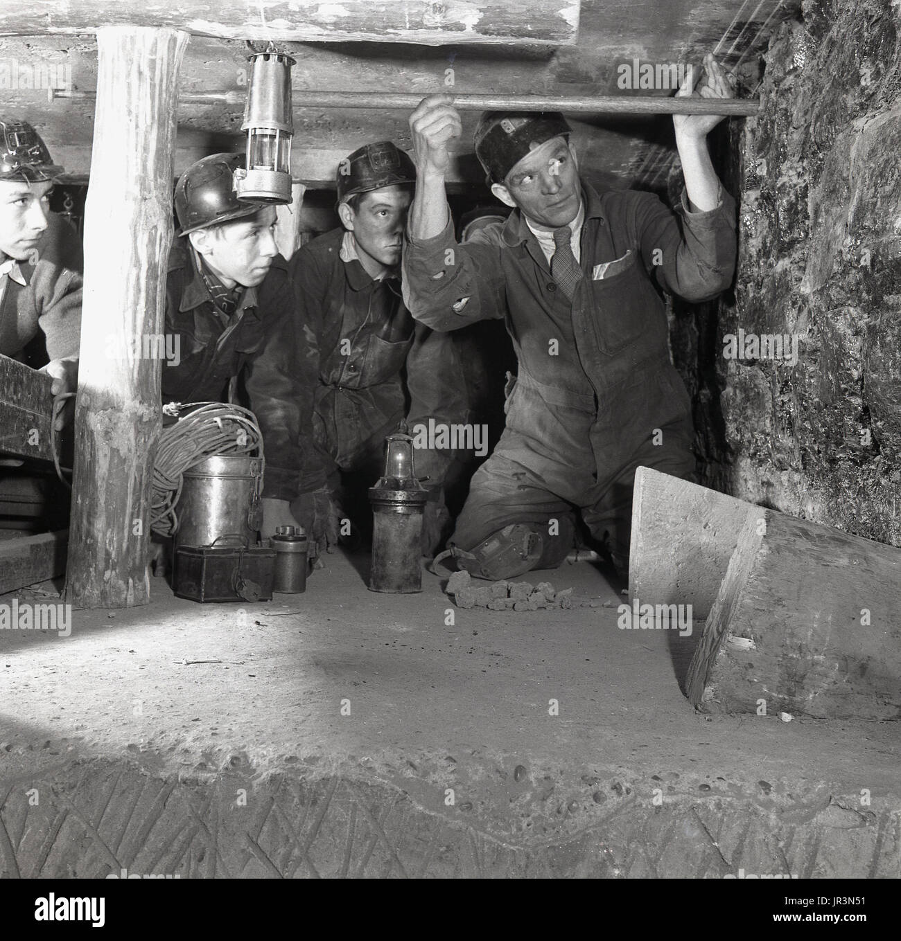 1948, storico, accovacciato in un pozzo sotterraneo di miniera, un supervisore minerario che dimostra ad alcuni apprendisti una tecnica mineraria, utilizzando un palo di legno per controllare la parete della miniera. Foto Stock