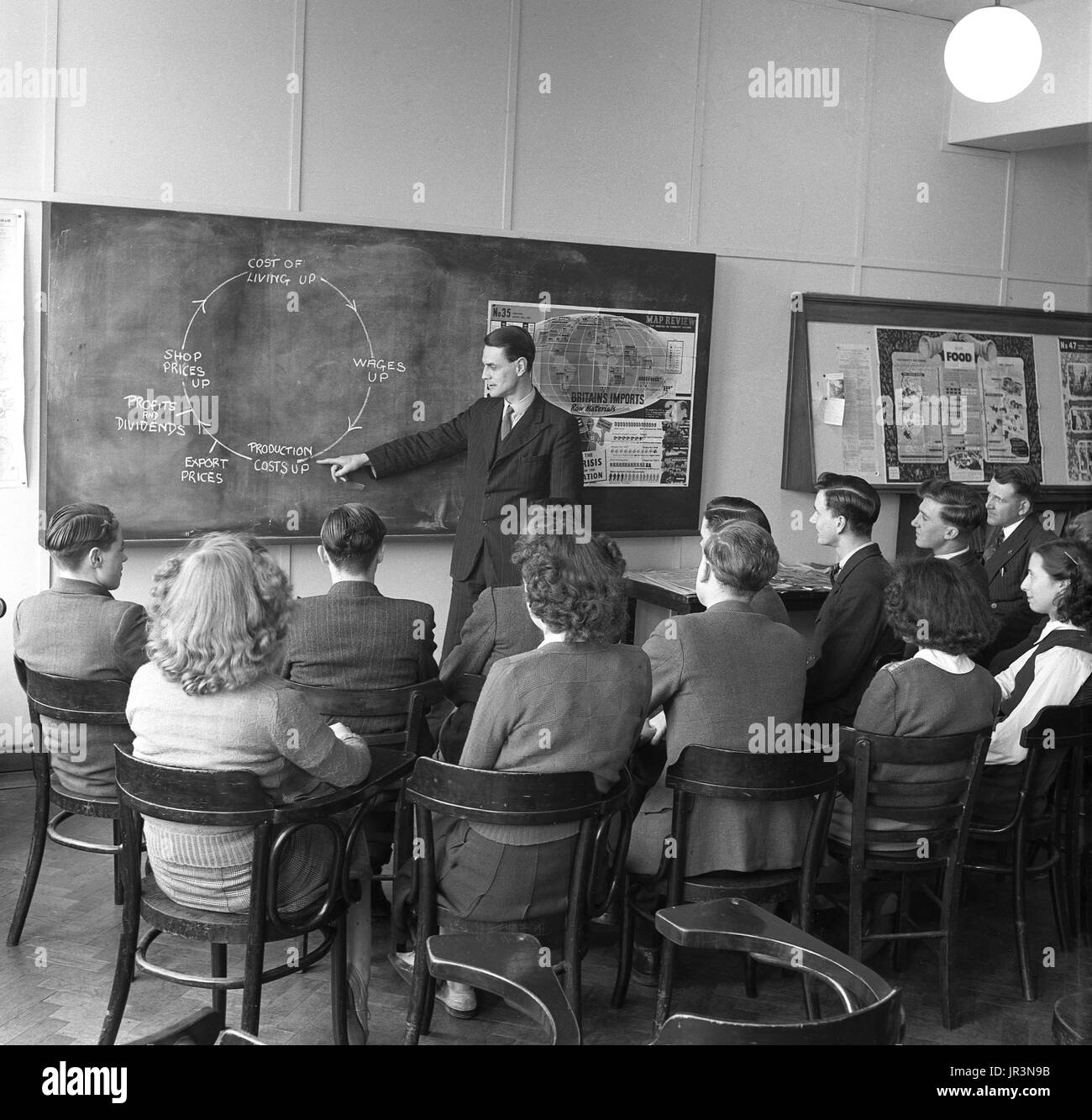 1948, storico, post-ww2 Gran Bretagna, eonomics lezione, un insegnante maschio utilizzando un diagramma sulla lavagna a spiegare a un gruppo di studenti maschi e femmine come il costo della vita è influenzata dalla Gran Bretagna alla necessità di importazione di materie prime. Foto Stock