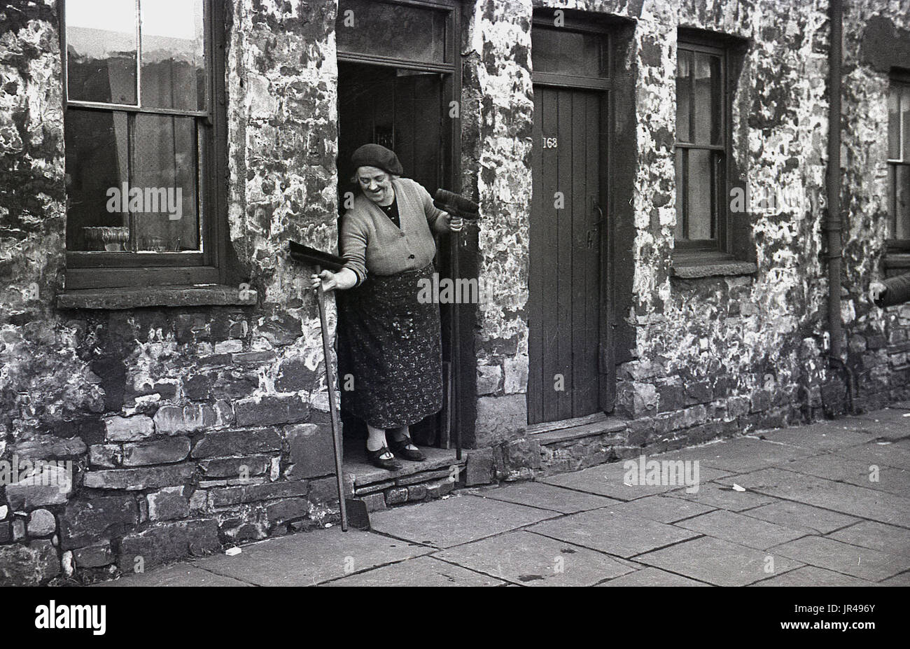 1940s, Merthyr Tydfil, South Wales, Regno Unito, signora anziana in grembiule con scope presso lo sportello anteriore della sua pietra costruito cottage. Foto Stock
