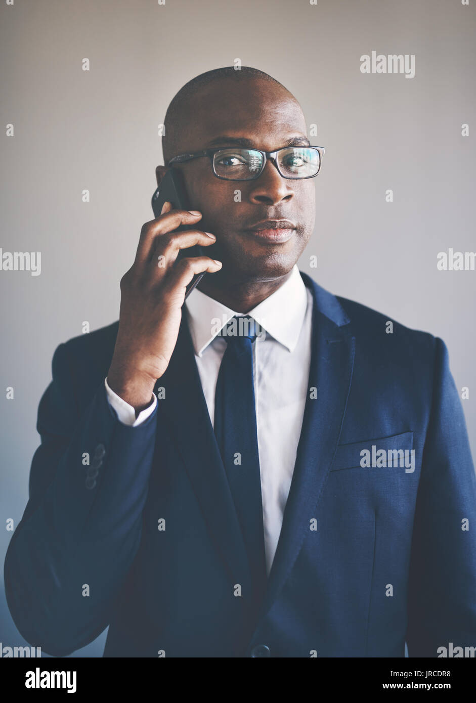 Ritratto di un fiducioso giovane imprenditore africano indossa una tuta e occhiali parlando al cellulare mentre sta in piedi da solo in un ufficio Foto Stock