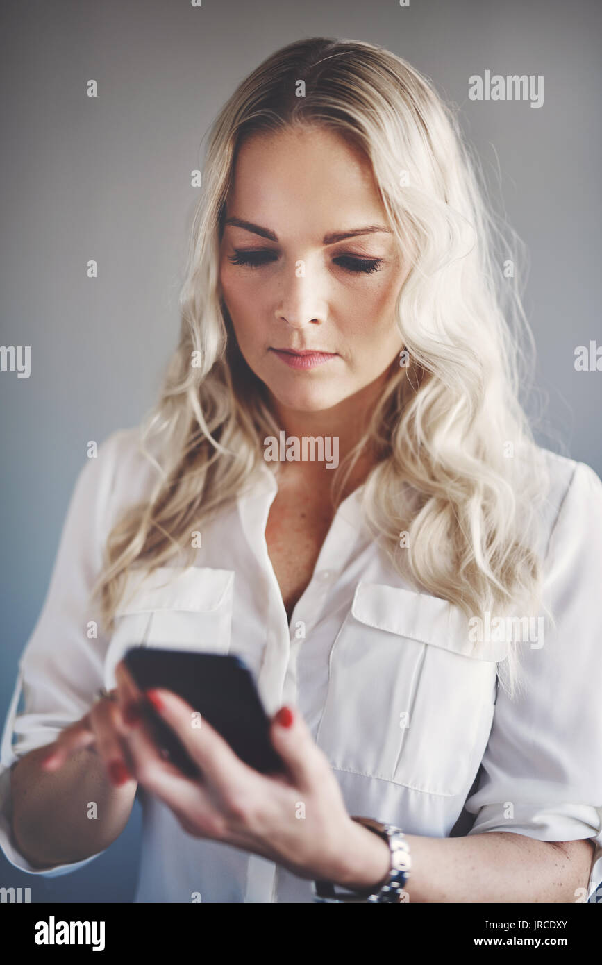 Giovane imprenditrice con lunghi capelli biondi di inviare messaggi di testo su un telefono cellulare mentre sta in piedi da solo in un ufficio Foto Stock