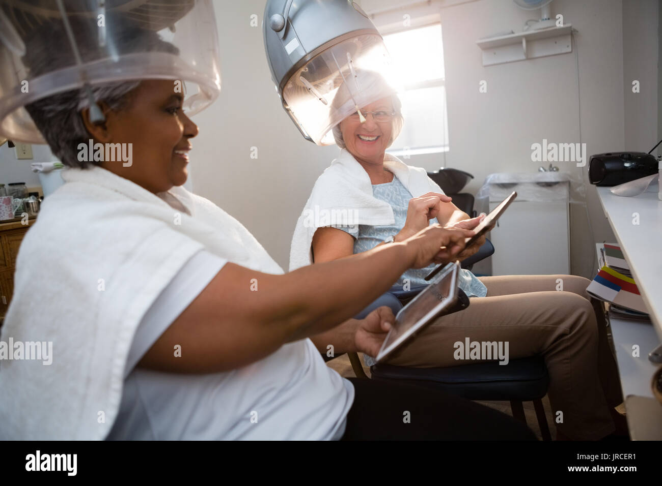 Amici utilizzando la tavoletta digitale mentre seduto sotto i capelli vapore a salon Foto Stock