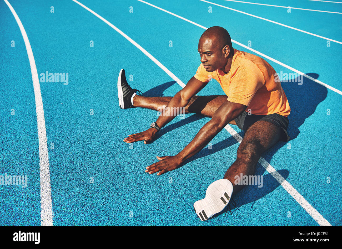 Concentrato di giovani africani atleta maschio in sportswear seduto da solo sulle corsie di una via di corsa stretching prima di una corsa Foto Stock