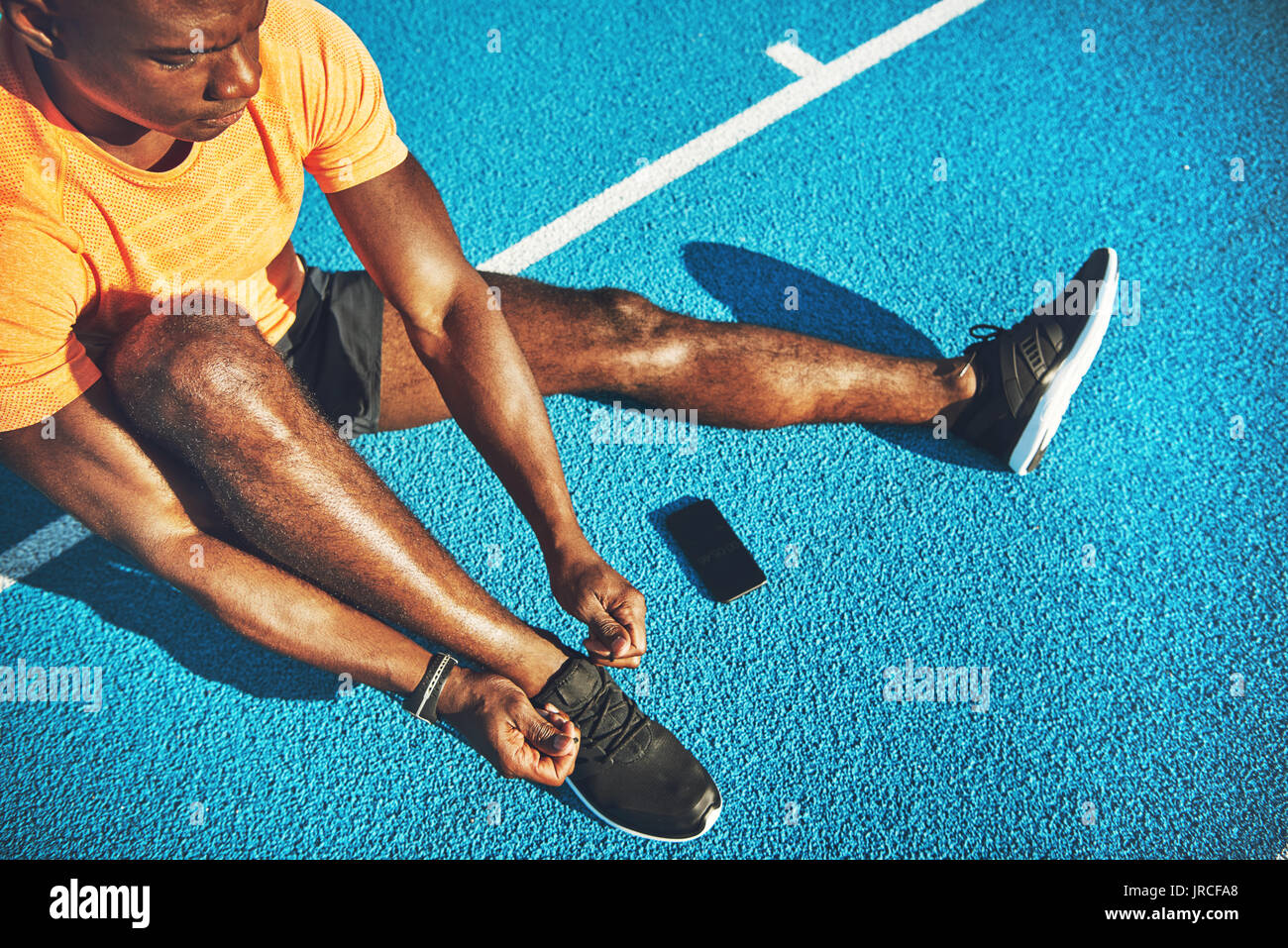 Concentrato di giovani africani atleta maschio in sportswear seduto da solo sulle corsie di una via di corsa occupare le sue scarpe prima di partecipare al corso di formazione Foto Stock