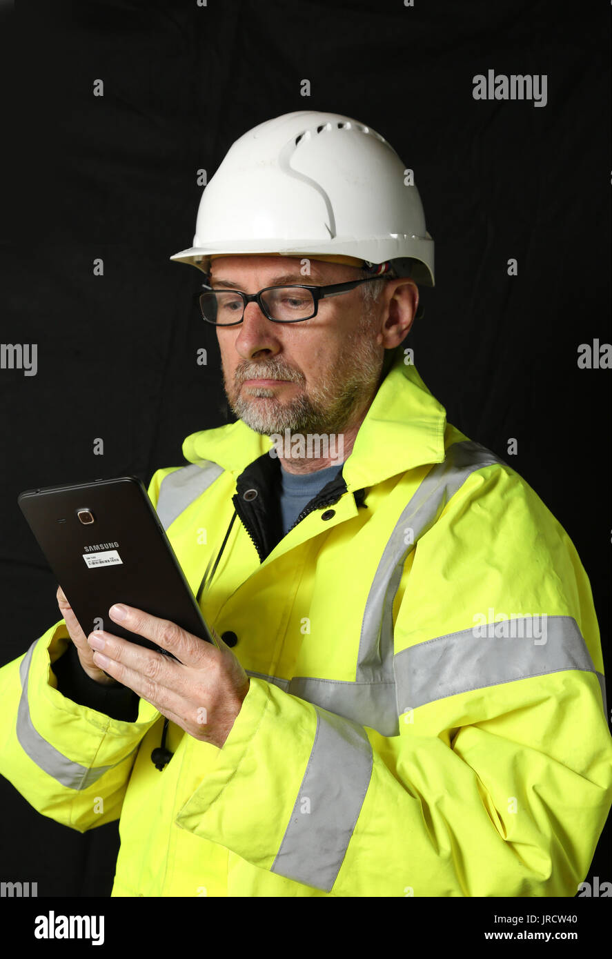 Un ingegnere edile con una giacca gialla ad alta visibilità studia un tablet. Sfondo chiaro e scuro. Foto Stock