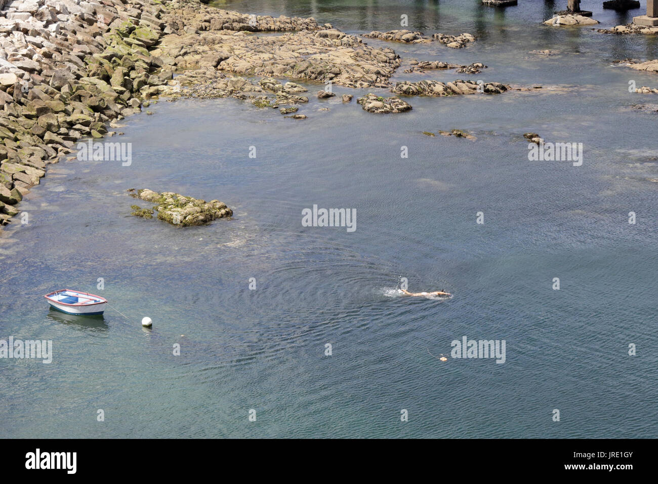 Un piccolo di legno bianco barca ancorata in un porto accanto a una roccia pier e scogliere con alcune boe e una piscina uomo Foto Stock