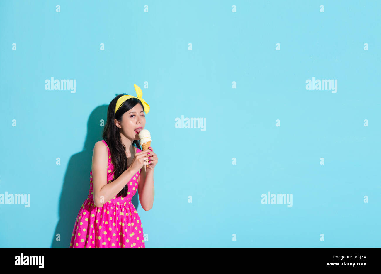 Bella donna elegante con una strizzatina d'occhio occhio che guarda la fotocamera e godendo litta deliziosi gelati in piedi su sfondo blu. Foto Stock