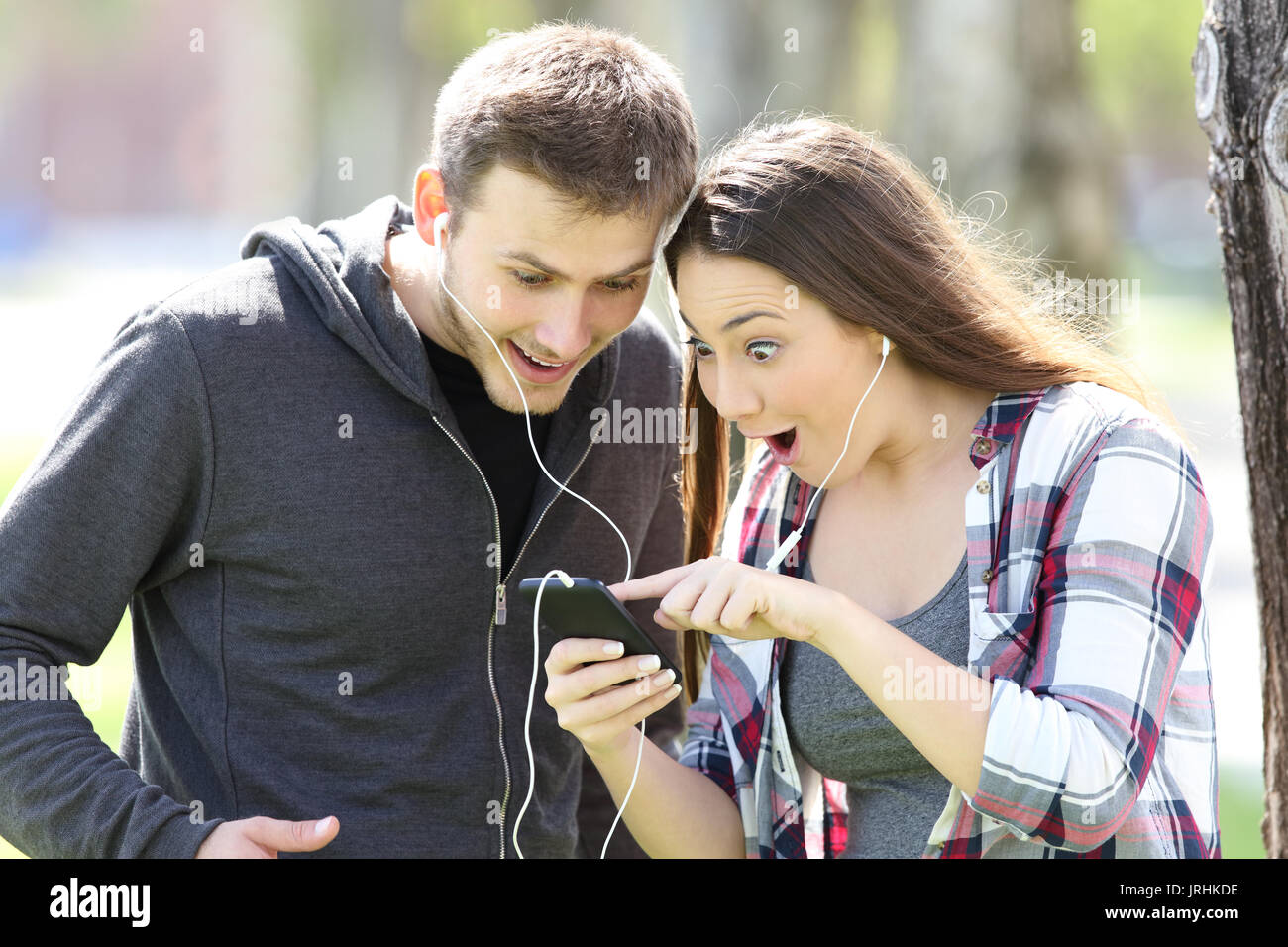 Stupiti giovane piedi ascoltando la musica on line e la visione di contenuti multimediali in uno smart phone all'aperto in un parco Foto Stock