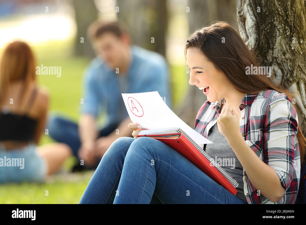 Studente eccitate controllando un esame approvato seduta sul prato in un parco con persone non focalizzato in background Foto Stock