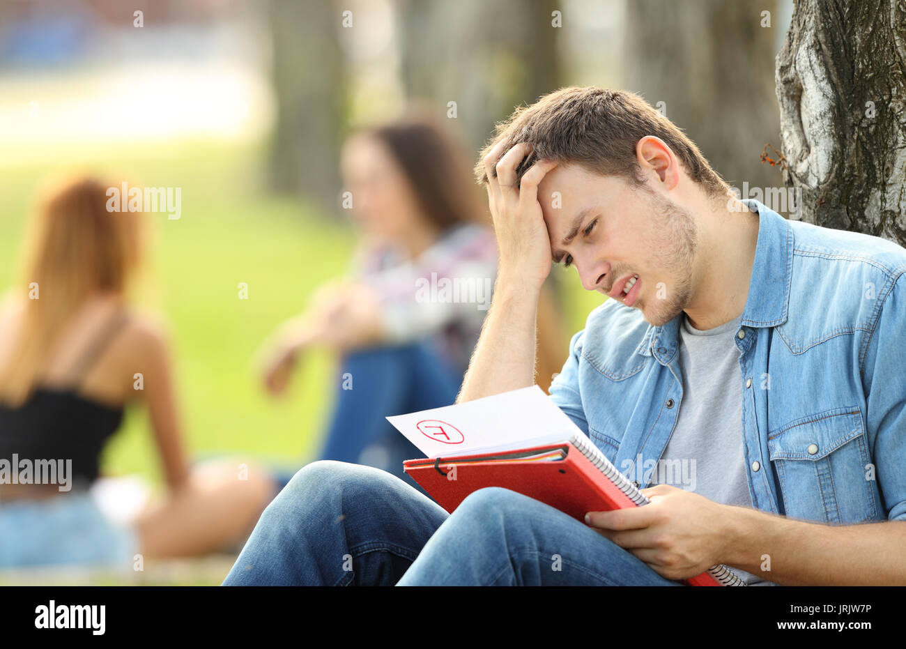 Singolo studente triste la verifica di un esame non superato seduto sull'erba in un parco con persone non focalizzato in background Foto Stock