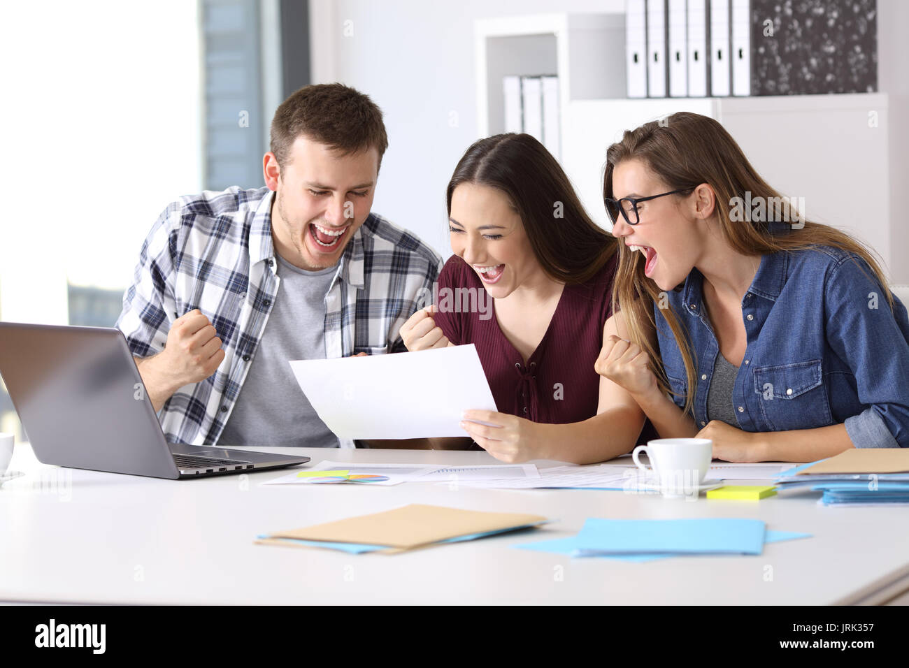 Tre lavoratori eccitato leggendo un buon rapporto risultati seduto in un desktop in ufficio Foto Stock