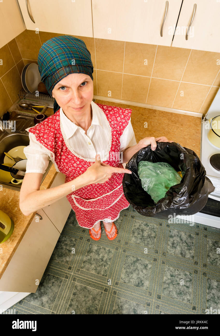 Una donna con una sciarpa sulla testa e un grembiule rosso, è guardando all'interno di un nero cestino con un sacco di immondizia in cucina. Lei è molto disturbato da th Foto Stock