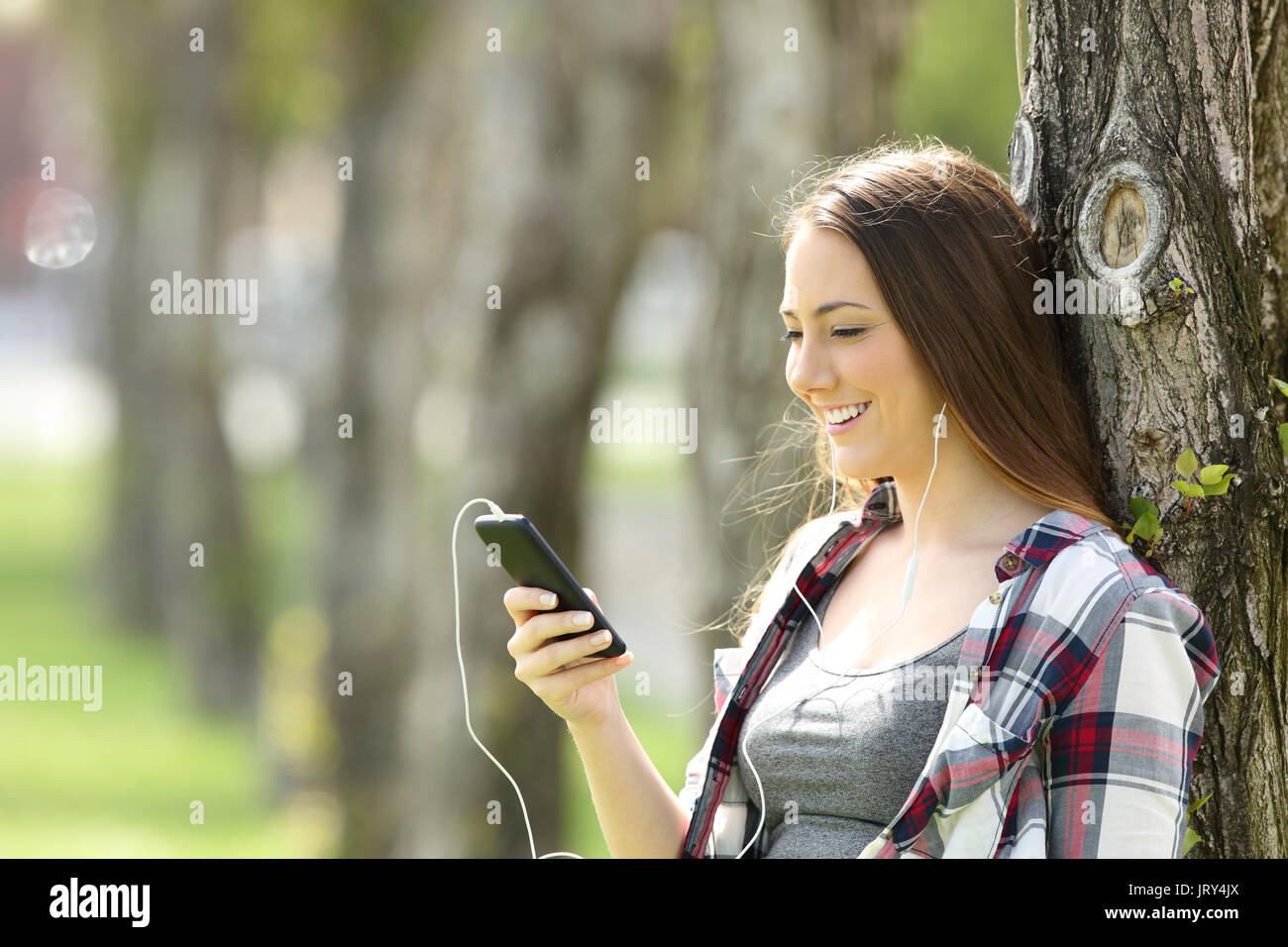 Felice teen ascoltare musica on line e la visione di contenuti in streaming in uno smart phone appoggiato su di un albero esterno Foto Stock