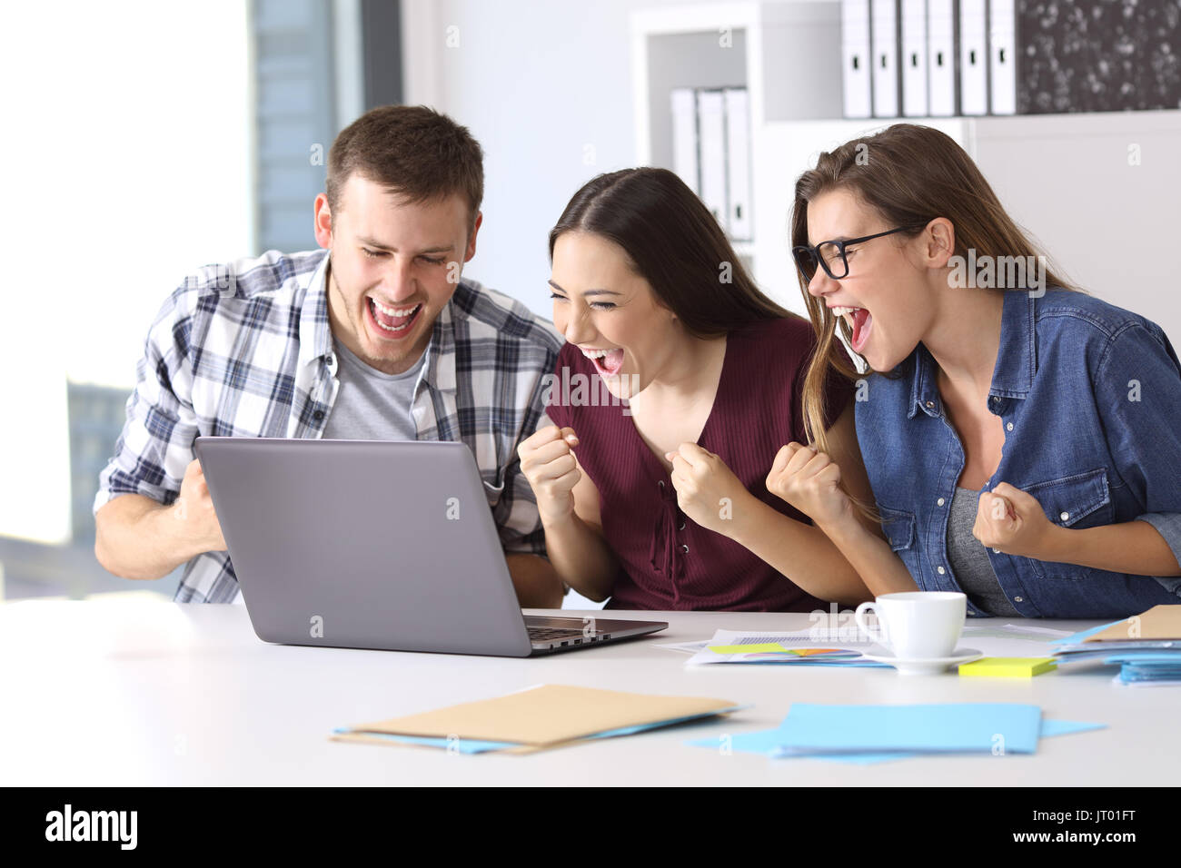 Tre dipendenti eccitato la lettura di buone notizie on line in una seduta del notebook in un desktop in ufficio Foto Stock