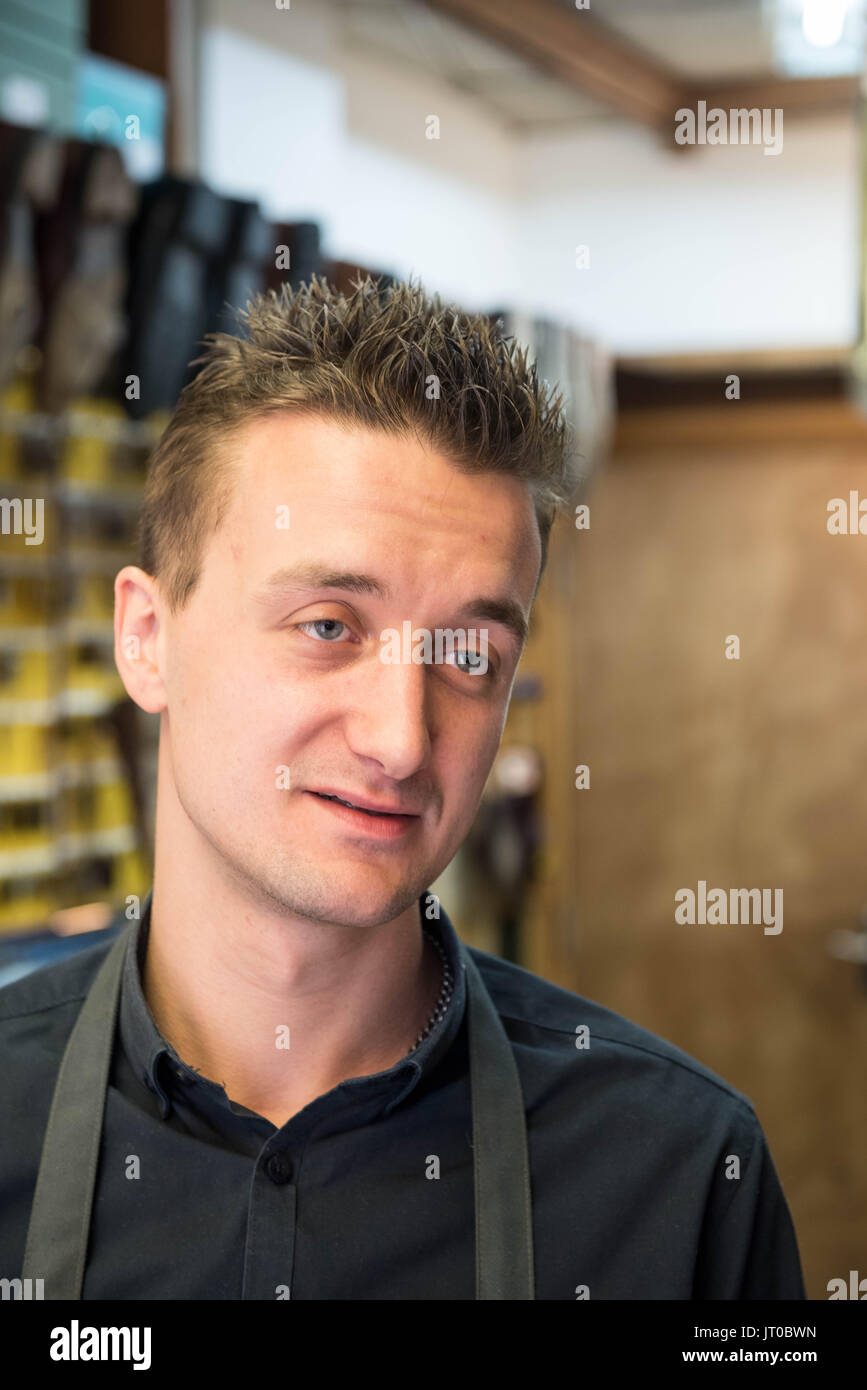 Cobbler che ripara scarpe in pelle all'interno di un'officina tradizionale. Sulla parete sono visibili utensili, attrezzature di taglio chiave e suole di ricambio Foto Stock