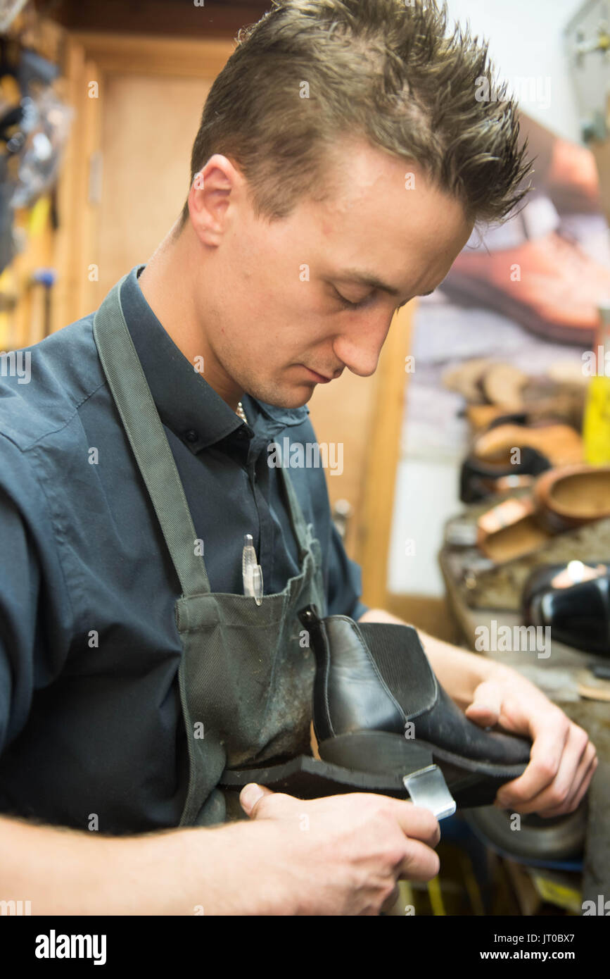 Cobbler che ripara scarpe in pelle all'interno di un'officina tradizionale. Sulla parete sono visibili utensili, attrezzature di taglio chiave e suole di ricambio Foto Stock