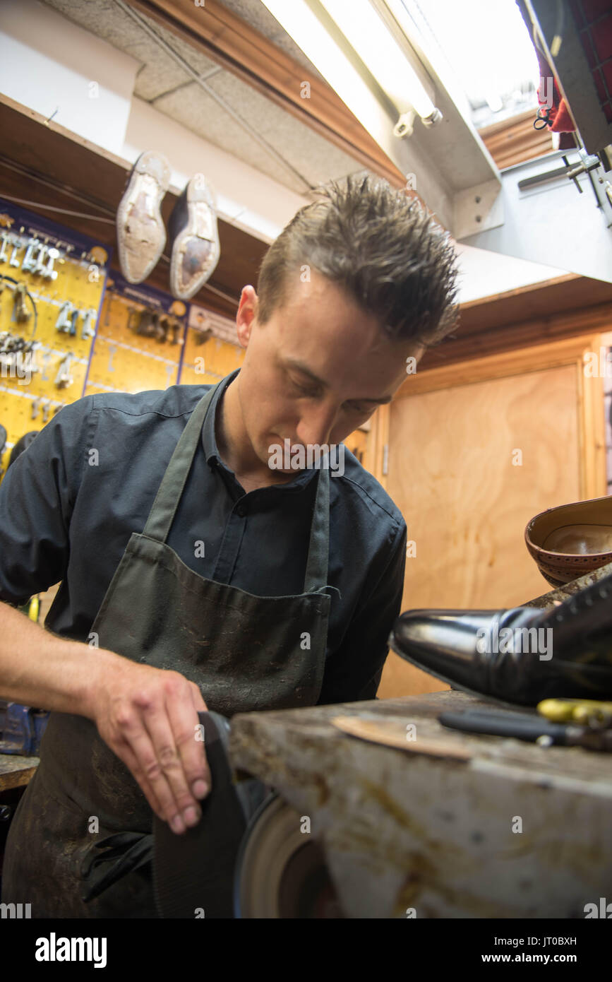Cobbler che ripara scarpe in pelle all'interno di un'officina tradizionale. Sulla parete sono visibili utensili, attrezzature di taglio chiave e suole di ricambio Foto Stock