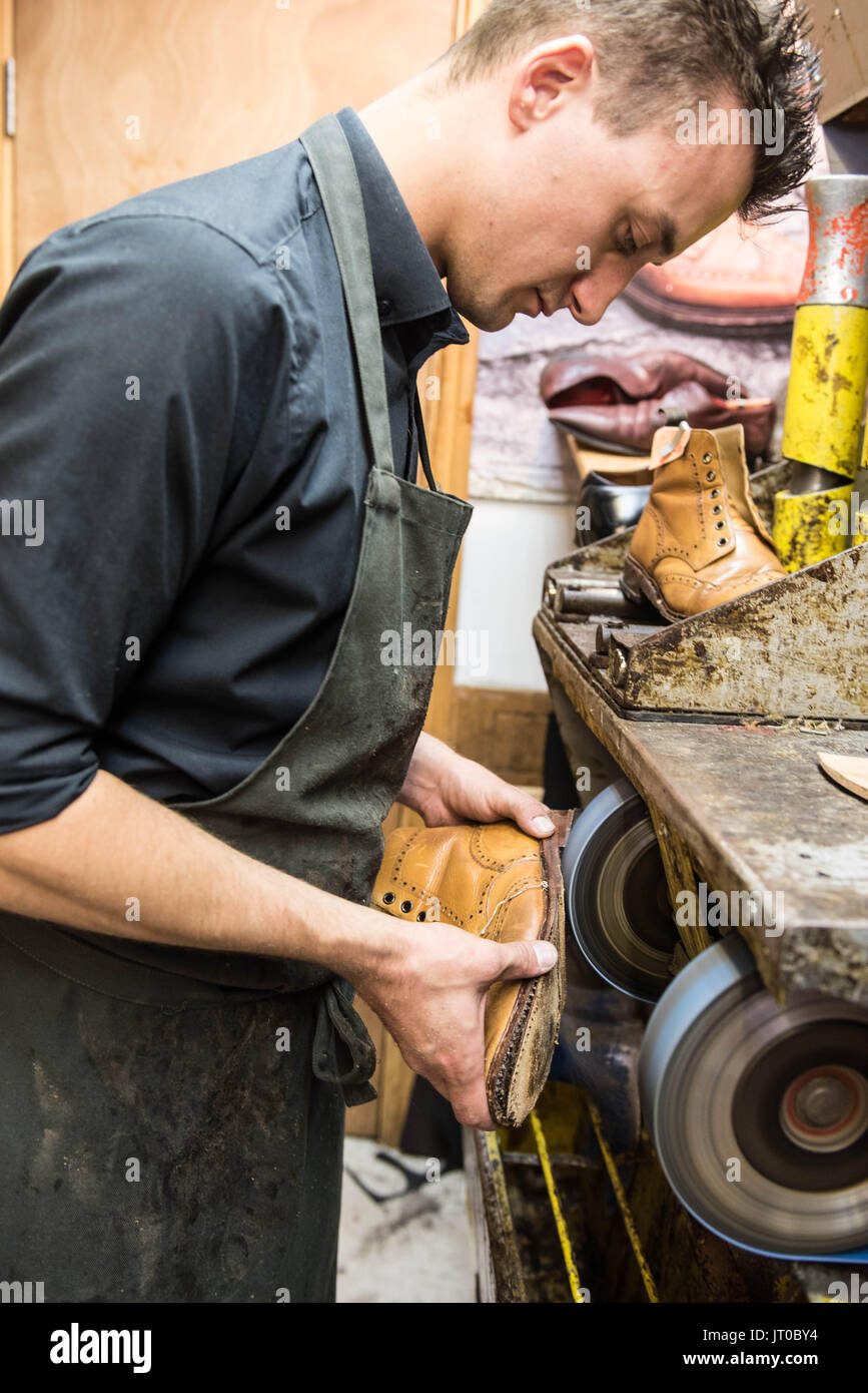 Cobbler che ripara scarpe in pelle all'interno di un'officina tradizionale. Sulla parete sono visibili utensili, attrezzature di taglio chiave e suole di ricambio Foto Stock