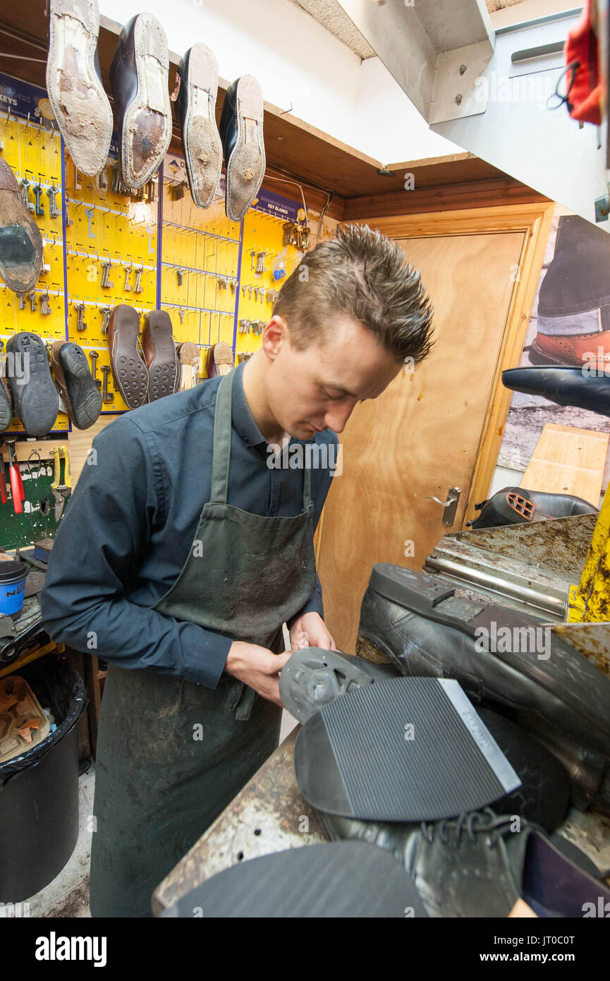 Cobbler che ripara scarpe in pelle all'interno di un'officina tradizionale. Sulla parete sono visibili utensili, attrezzature di taglio chiave e suole di ricambio Foto Stock