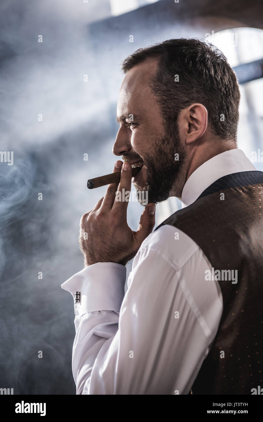 Vista laterale di sorridere fiducioso uomo sigaro fumare in luoghi chiusi Foto Stock